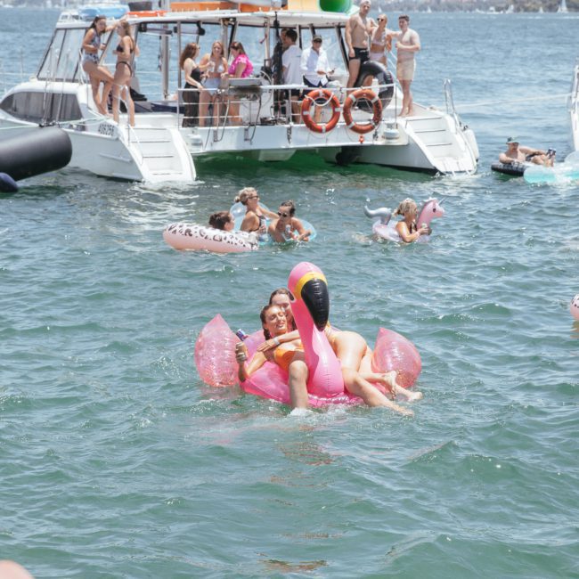 People enjoying a sunny day swimming and floating on inflatables in the water near several boats. Two people are prominently riding a pink flamingo inflatable by a luxury yacht hire Sydney.