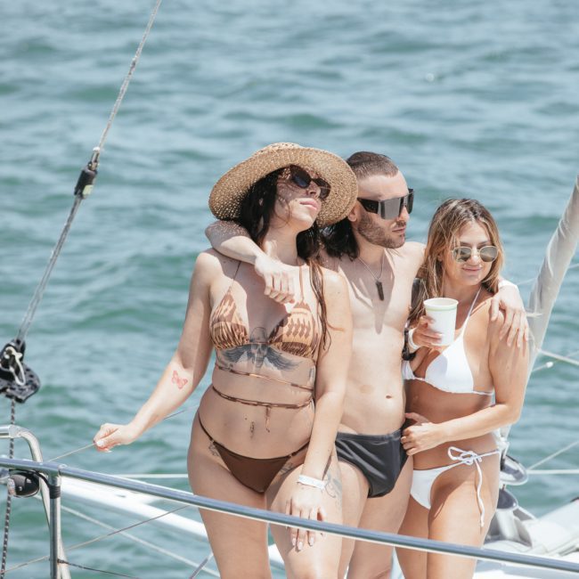 Three people in swimwear, standing close together on a sailboat, with an ocean backdrop. One person wearing a sunhat and bikini, another in swim trunks, and the third holding a cup. Enjoying the perfect vibe of a Sydney boat party hire!