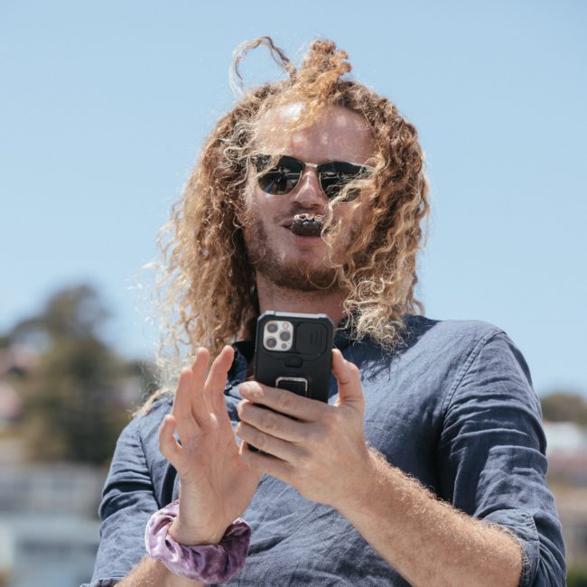 A person with long curly hair and sunglasses smiles while holding a smartphone outdoors, with buildings and trees in the blurred background, enjoying thoughts of a private yacht charter Sydney Harbour.