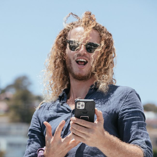 A man with curly hair and sunglasses smiles while holding a smartphone, with a blurred outdoor background featuring buildings and trees, likely planning his next adventure on a private yacht charter Sydney Harbour.