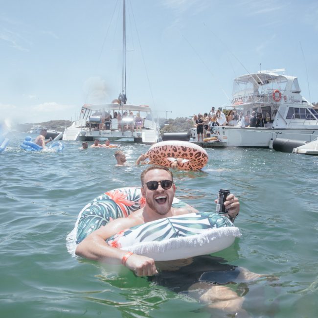 A person floats in the water with a beverage, surrounded by inflatable tubes and boats filled with people under a clear sky, enjoying the vibes of a Sydney boat party hire.