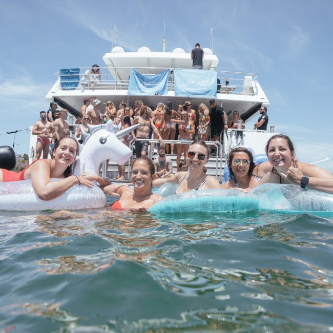 A group of five people float on inflatable toys in the water near a private yacht charter in Sydney Harbour, set against a clear sky.