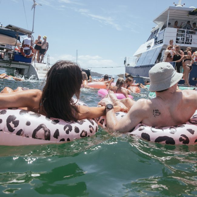 People in inflatable tubes relax in the water among several boats on a sunny day, with more individuals socializing and sunbathing on the decks of the nearby vessels, enjoying what looks like a lively Catamaran party Sydney.