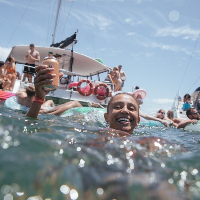 A person in the water holds a drink and smiles at the camera while others relax on floats and a boat in the background, enjoying a corporate boat event in Sydney.