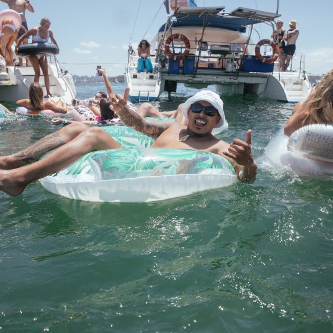 Person lounging on an inflatable float in the water, surrounded by others on similar floats and boats in the background on a sunny day. It's like a mini Catamaran party Sydney style!