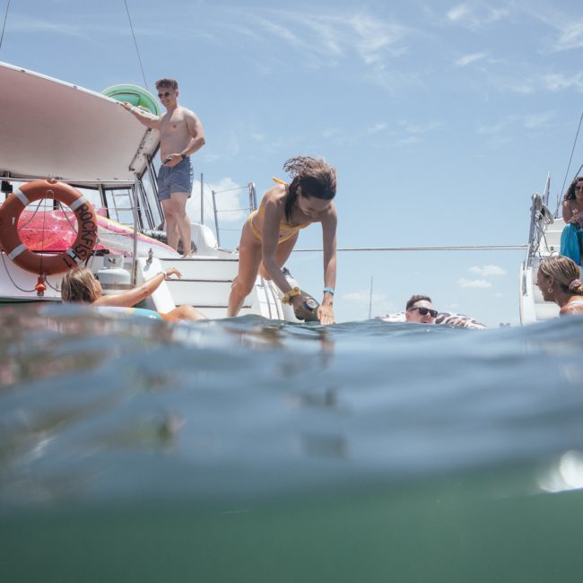 People are interacting on and around luxury yachts in a body of water on a sunny day, some are swimming while others remain on the boats enjoying the clear blue sky.