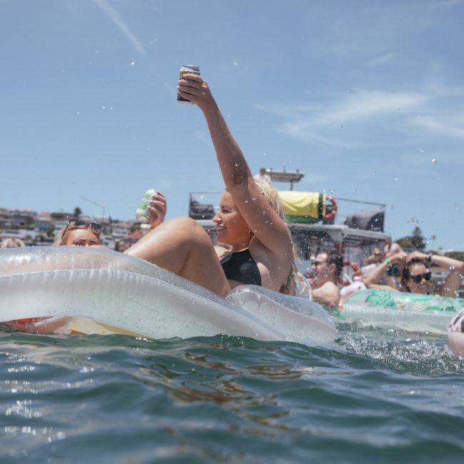 People are relaxing on inflatable rafts in a body of water, holding drinks, with boats and a shoreline visible in the background. Nearby, a DJ boat hire Sydney adds lively tunes to the vibrant scene of fun and leisure.