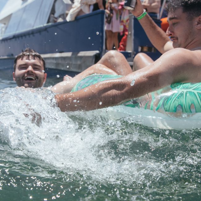 Two men in swim trunks enjoy floating in the water on inflatable tubes near a boat, with one splashing water playfully. They seem to be having a great time during their luxury yacht hire Sydney adventure.