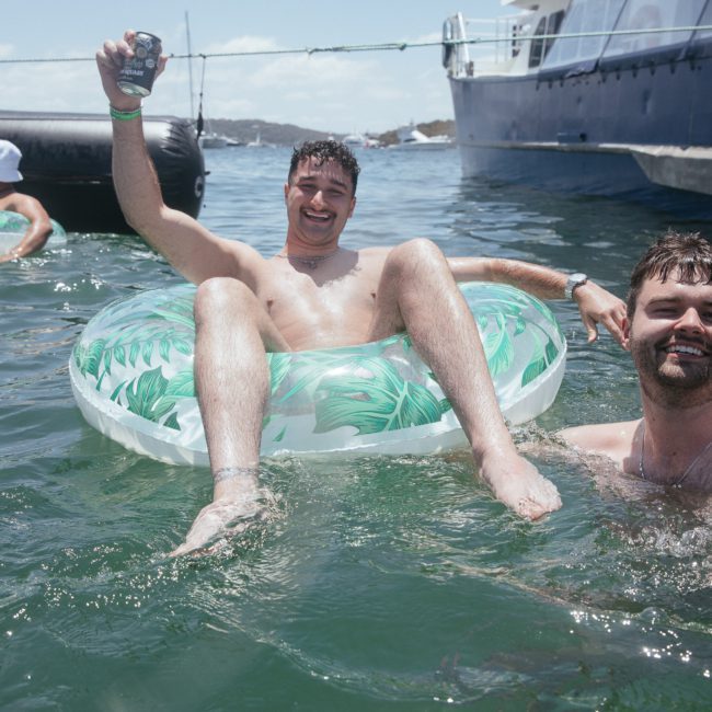 Two men enjoying time in the water; one is floating on an inflatable ring holding a drink and smiling, the other is partly submerged, smiling, and holding onto the inflatable. A private yacht charter cruises nearby, adding a luxurious touch to their fun.