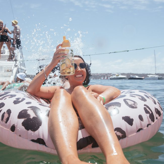 A person lounges on a leopard-print inflatable tube in the water, holding a drink bottle while others enjoy a private yacht charter in Sydney Harbour.