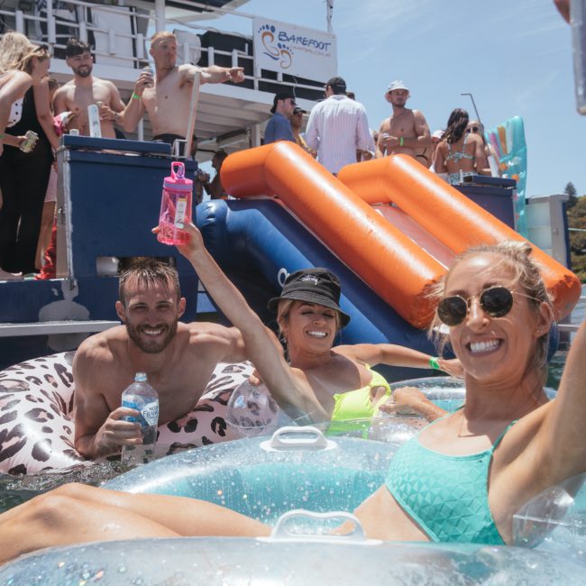 Three people in swimsuits lounge on floaties, raising drinks, in front of a luxury yacht hire Sydney with multiple partygoers and water slides. It's a sunny day.