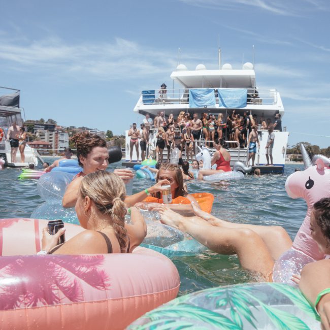 People on inflatable floats in the water near a boat. A group on the boat watches, while individuals on the floats appear to be relaxing and socializing. The sky is clear, and the setting is sunny—a perfect day for a catamaran party Sydney.