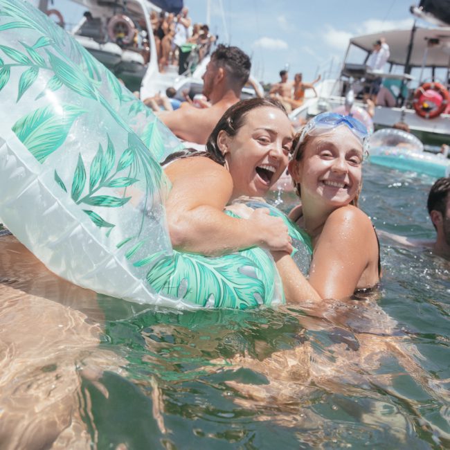 People swimming and enjoying themselves in the water, some using inflatable rings, with boats in the background on a sunny day. Perfect for those considering DJ boat hire Sydney or Corporate boat events Sydney for their next celebration.