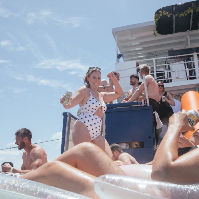 A woman in a white polka dot swimsuit stands on a boat deck holding a drink, while other people relax and enjoy drinks on inflatable pool rafts under a sunny sky. Experience the ultimate fun with Sydney boat party hire for an unforgettable day!