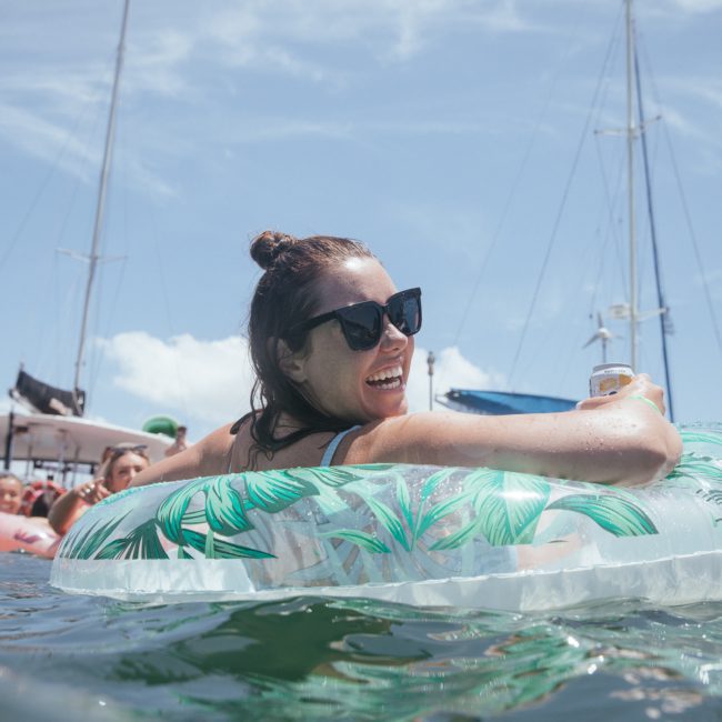 A person wearing sunglasses and a swimsuit floats on an inflatable ring in the water, surrounded by others and boats under a sunny sky, enjoying a lively DJ boat hire Sydney event.