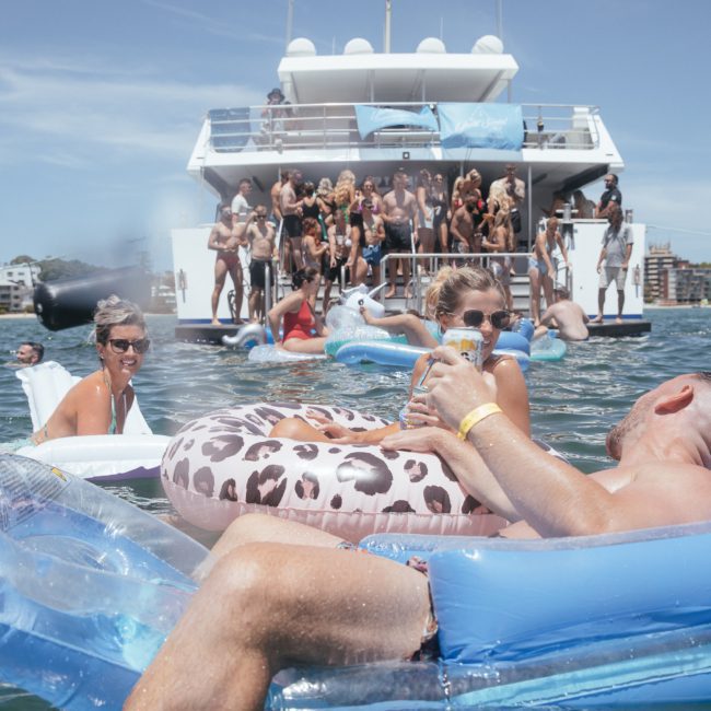 People relaxing in inflatable rings in the water while others gather on the back of a large yacht, perfect for corporate boat events Sydney.