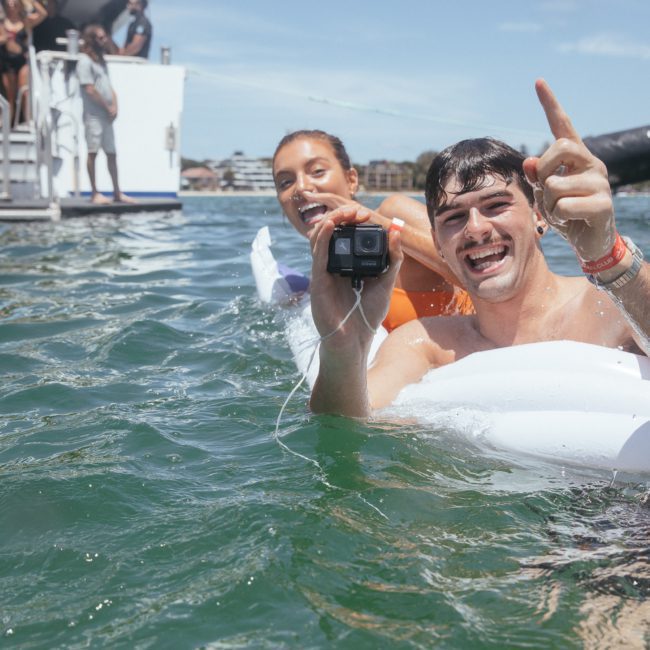 Two people in swimsuits float on inflatable rings in the water, holding a camera and smiling. More people are visible on a boat and stairs in the background under the sunny sky, enjoying their Sydney boat party hire.