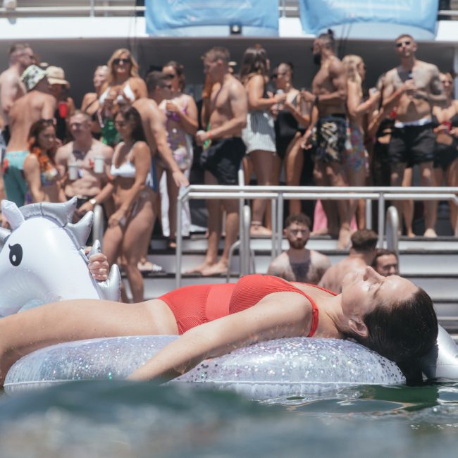 A group of people enjoying a catamaran party in Sydney while a woman in a red swimsuit lounges on a unicorn float in the water.