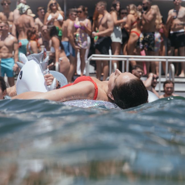 A crowd of people in swimwear is gathered on a boat and in the water, with some on inflatables. One person in a red swimsuit lies on an inflatable unicorn in the foreground, embodying the vibrant spirit of a Sydney boat party hire.