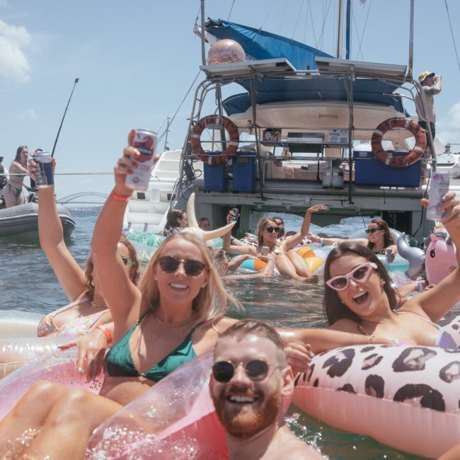 A group of people enjoying drinks and lounging on inflatable rings in the water near anchored boats on a sunny day, perfect for a private yacht charter Sydney Harbour.