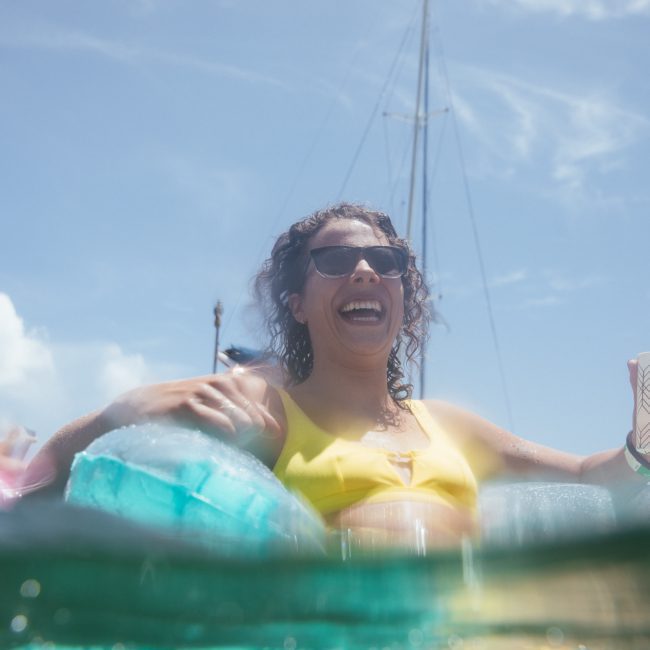 A woman in sunglasses and a yellow swimsuit laughs while holding a cup, half-submerged in water with yachts, enjoying a private yacht charter on Sydney Harbour in the background.