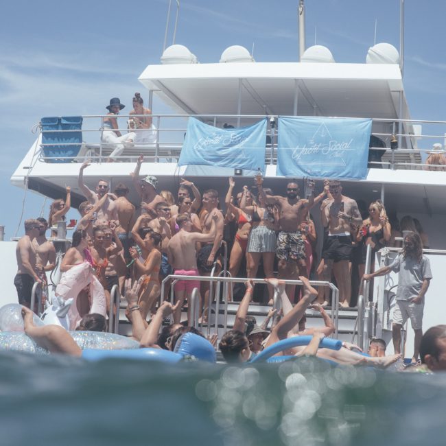 A group of people in swimwear are on and around a docked boat, some in the water, and many posing and waving. Blue banners with the text "White Sand" are on the boat's upper deck railing, creating a lively atmosphere perfect for corporate boat events Sydney.