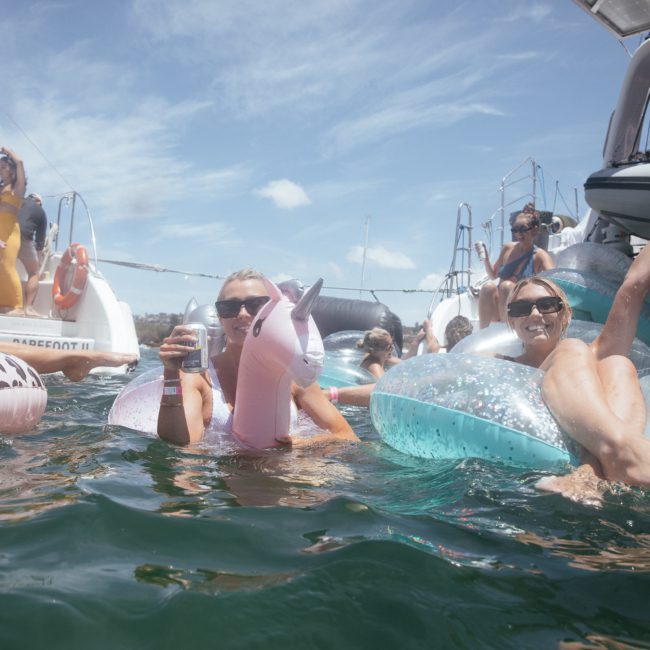 People floating in the water on inflatable pool toys, holding drinks, surrounded by boats on a sunny day at a Sydney boat party hire.