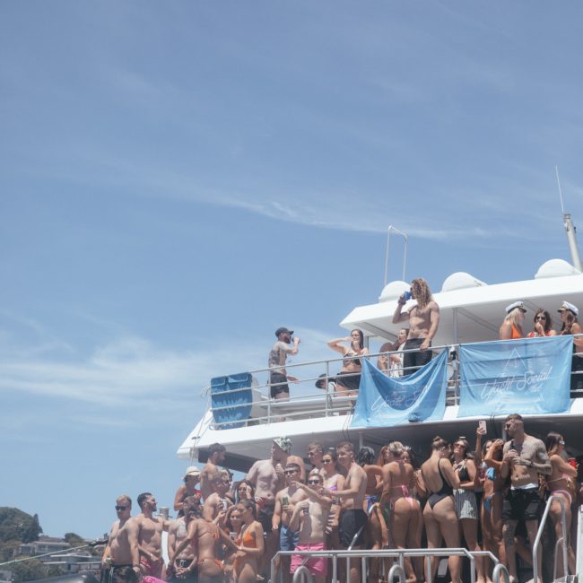 A large group of people in swimwear are gathered on multiple levels of a boat, enjoying blue skies and water in the background. Experience the excitement with a DJ boat hire Sydney for an unforgettable day out.