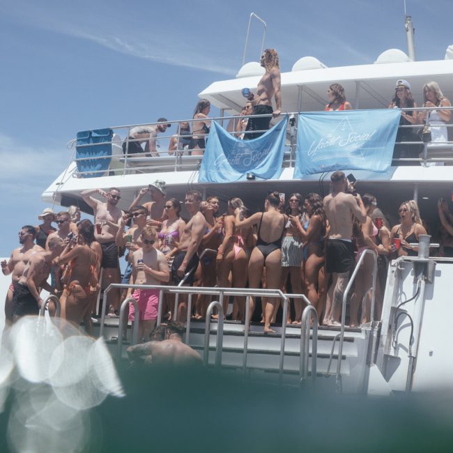 A large group of people in swimsuits stands on the deck of a boat under a clear sky, with some looking out towards the water and blue banners displaying "White & Case" visible in the background, enjoying what looks like a lively catamaran party Sydney.