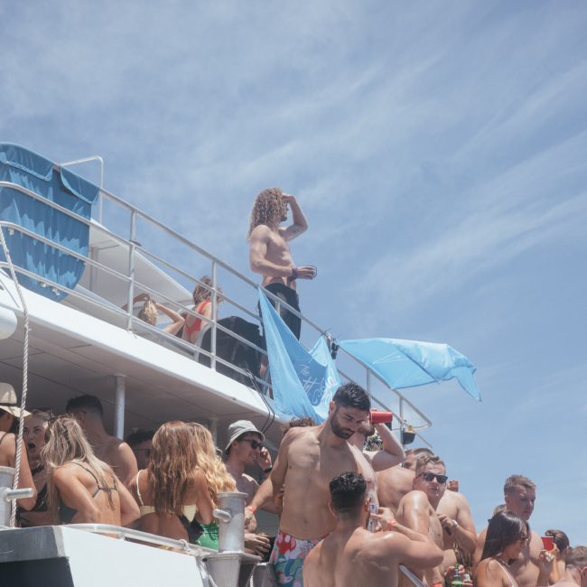 A group of people, some in swimwear, are gathered on the deck of a boat under sunny skies. One person stands on the upper level, holding onto a blue flag. Enjoying their time on a luxury yacht hire in Sydney Harbour.