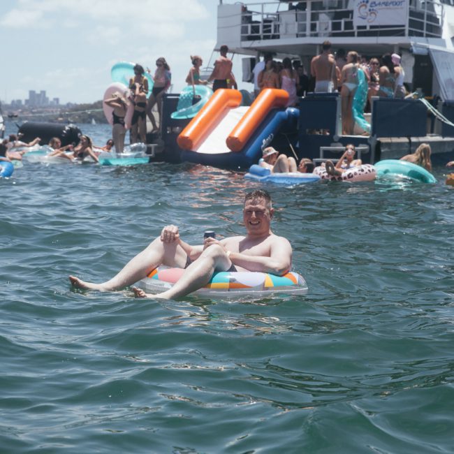 Person on a colorful inner tube in the water with a crowded boat and other floating devices in the background on a sunny day, enjoying a catamaran party Sydney style.