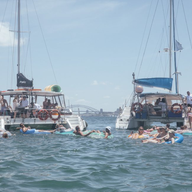 A group of people relax on inflatables in the water between two anchored sailboats on a sunny day, with a cityscape and bridge visible in the background, enjoying a luxury yacht hire Sydney experience.
