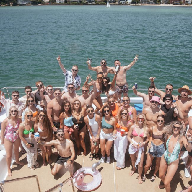 A group of people on a boat posing for a photo. Some are standing, others are seated, and many are holding drinks. They are dressed in swimwear and casual summer clothing, enjoying a luxury yacht hire Sydney. The water and shoreline are visible in the background.