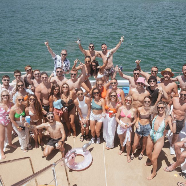 A large group of people in swimwear posing and celebrating on the deck of a boat, surrounded by water on a sunny day, epitomizes the vibrant atmosphere you can expect with Sydney boat party hire.
