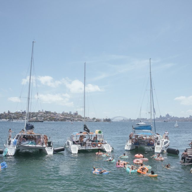 Four catamarans are anchored in the water with people swimming and floating on inflatables, enjoying a vibrant catamaran party Sydney style. The stunning Sydney cityscape and iconic Sydney Harbour Bridge are visible in the background.