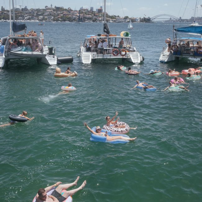 Groups of people on inflatable floats in the water between boats in a bay, with city buildings and a bridge in the background, enjoying the sights during luxury yacht hire Sydney.