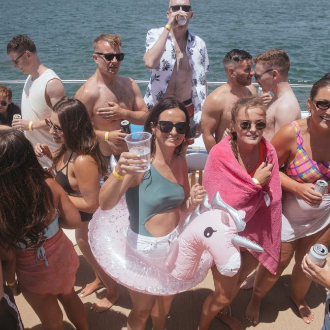 A group of people, some in swimwear and flotation devices, enjoy drinks on a luxury yacht under sunny weather. A woman in a unicorn float and sunglasses raises her glass. Perfect for a private yacht charter on Sydney Harbour.