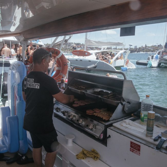 A person cooks meat on a grill aboard a private yacht charter in Sydney Harbour. Other boats and people are seen in the water and in the background, under sunny weather. An inflatable unicorn lies nearby.