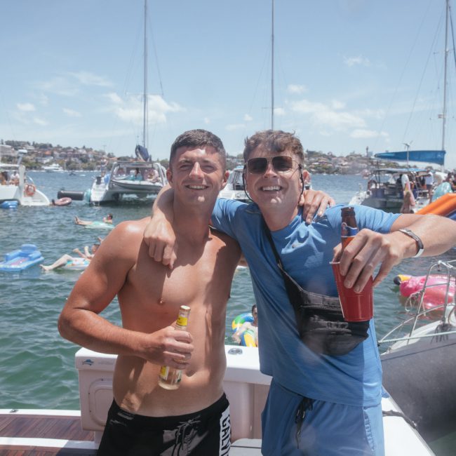Two men smiling with arms around each other on a boat, holding drinks, with other people, boats, and a scenic background of water and buildings. Sydney boat party hire offers unforgettable experiences like these for friends and colleagues.