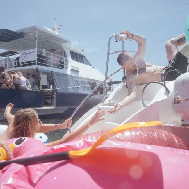 People enjoying a sunny day on the water with inflatables near a docked boat, while one person reaches in an attempt to climb onto a floating inflatable unicorn. Consider this scene for luxury yacht hire Sydney or corporate boat events Sydney for your next unforgettable outing.