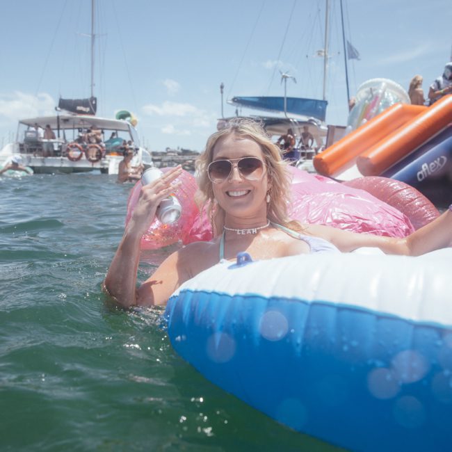 A woman is floating on an inflatable in the water, holding a can in each hand. Boats and other people on inflatables are in the background under a sunny sky, enjoying what appears to be a lively catamaran party Sydney style.