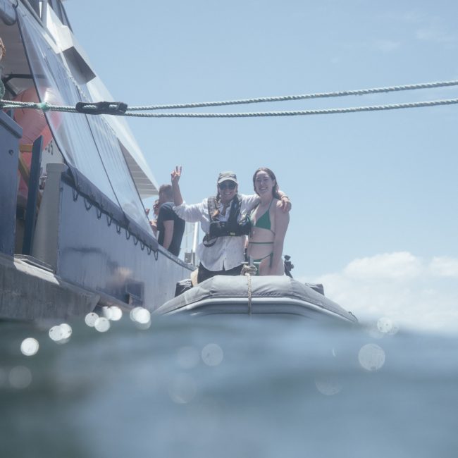 Two people on a small boat wave towards the camera. One person is wearing a headscarf and the other is in a swimsuit. A larger boat, indicative of luxury yacht hire Sydney, is anchored nearby. The sky is clear and the water calm, creating a perfect scene for relaxation on Sydney Harbour.