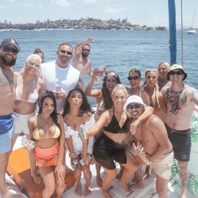 A group of people on a boat enjoying a sunny day on the water. Some are wearing swimsuits, sunglasses, and hats. They are posing for the camera with drinks and smiling against the stunning cityscape in the background —a perfect Catamaran party Sydney experience.