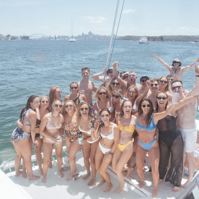 A group of people in swimwear poses for a photo on a catamaran party with a city skyline and water in the background.