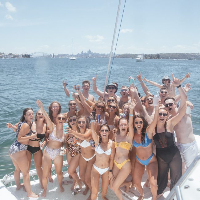 A group of people in swimwear poses on a private yacht charter in Sydney Harbour on a sunny day, with a cityscape and harbor in the background.