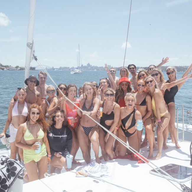 A group of people in swimwear stand closely together on a boat under sunny skies, smiling and holding drinks, with water and a cityscape in the background—a perfect scene from a Sydney boat party hire.