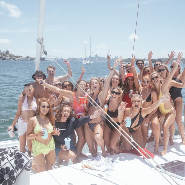A group of people in swimwear are posing and raising their arms excitedly on a catamaran party in Sydney Harbour on a sunny day, with a cityscape visible in the background.