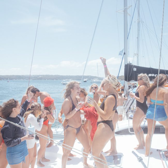 A group of people in swimwear are gathered on a private yacht charter in Sydney Harbour, enjoying drinks and socializing on a sunny day with other boats visible in the background.
