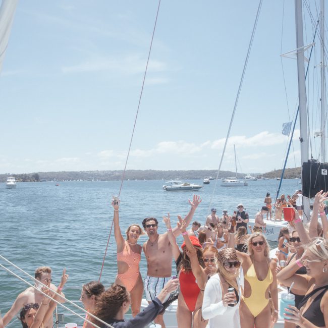 A group of people in swimwear are gathered on a luxury yacht hire Sydney, smiling and raising their arms in a celebratory manner. The boat is situated in a body of water with other boats and a shoreline visible in the background.