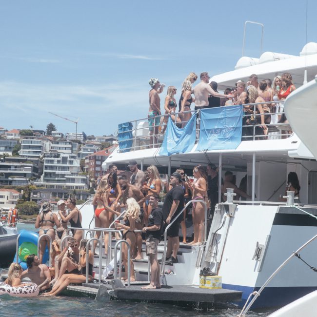 A group of people in swimwear gather and socialize on the deck of a luxury yacht hire Sydney in a sunny, coastal area with buildings in the background.